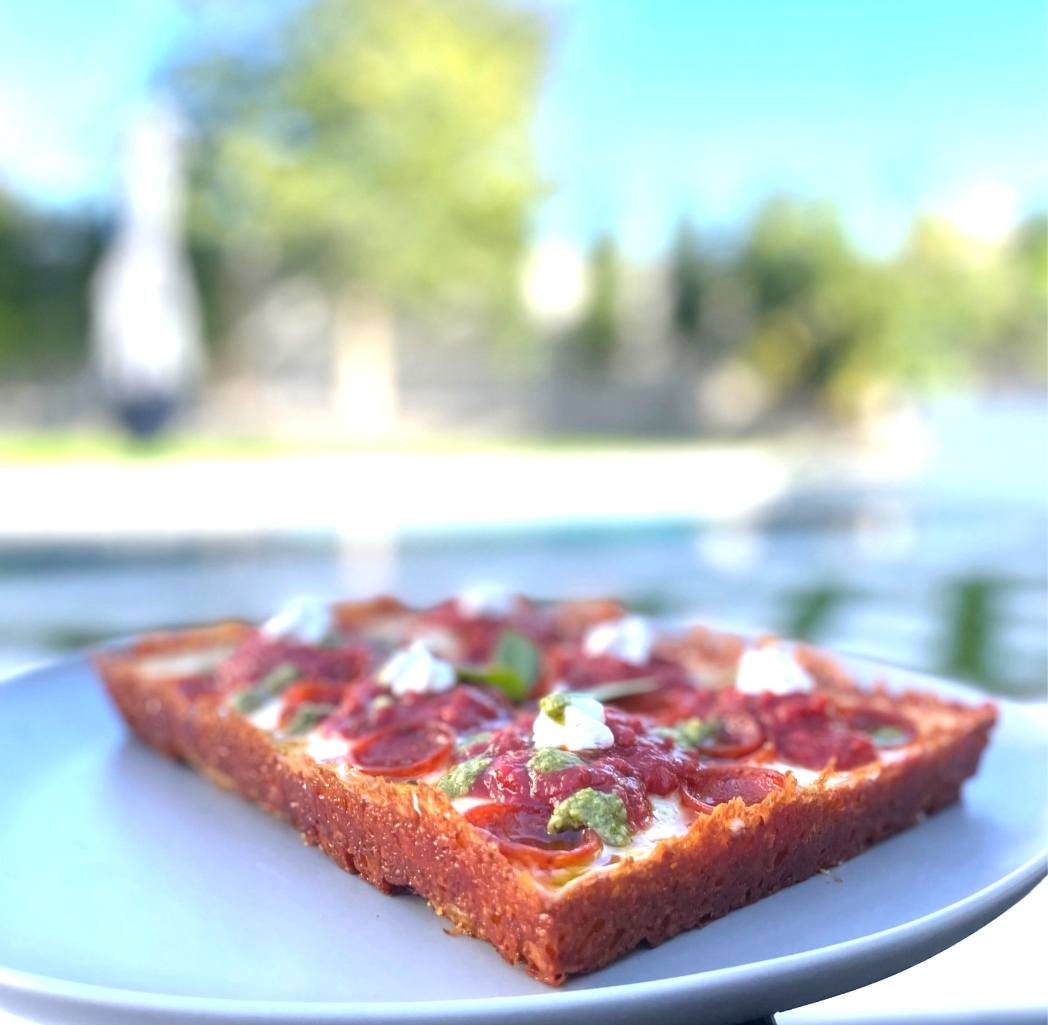 A Detroit-style slice on a blue plate with a backyard pool blurred behind it.