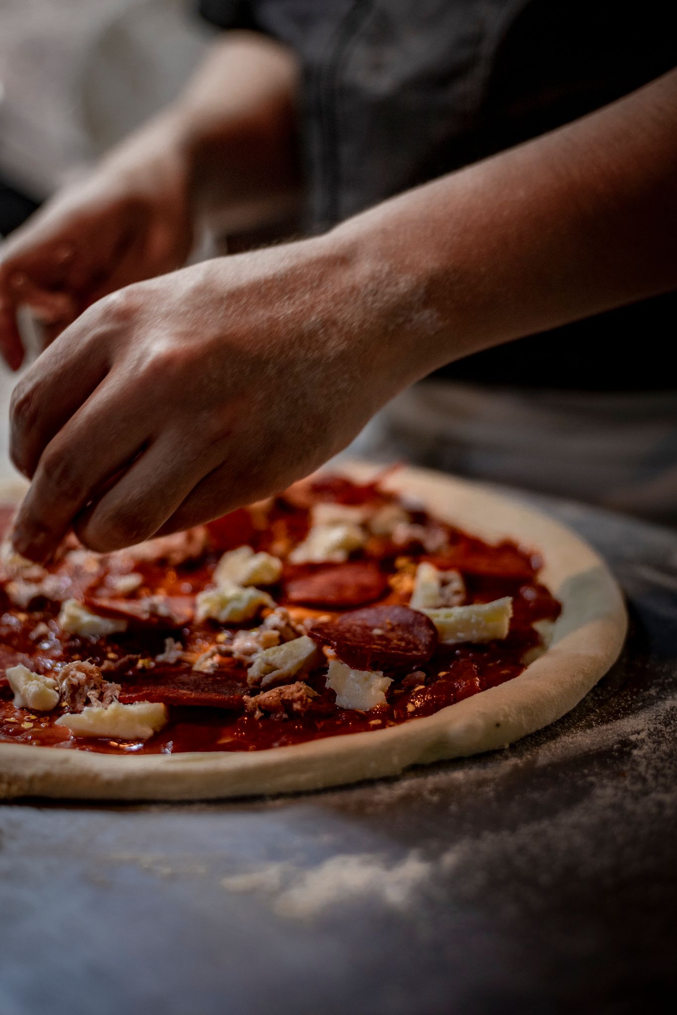 Hands of a chef skillfully preparing a pepperoni pizza with fresh ingredients in a kitchen setting.